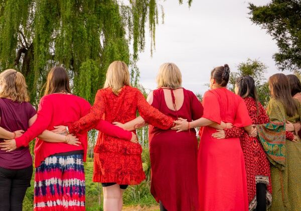 Women dressed in red, their backs to us, outside under a willow tree, with arms linked.